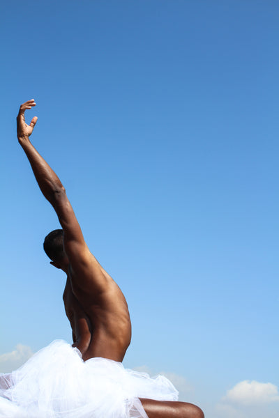 Black male dancing in the nature. His hand is oriented up onto the blue sky. We see him from behind. his armpit is glowing into the sunlight.